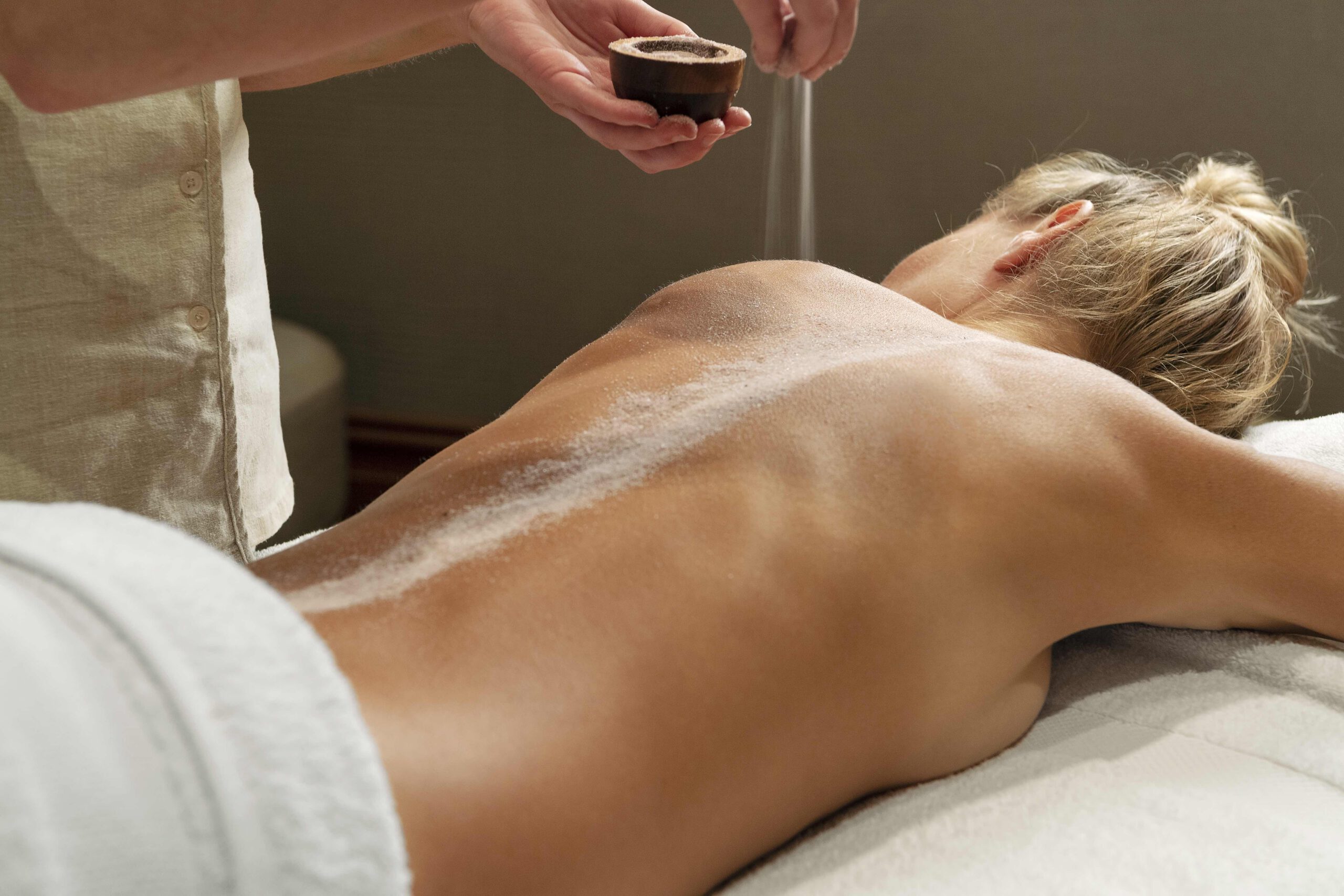 Woman lies on massage bed, therapist sprinkles material on her back